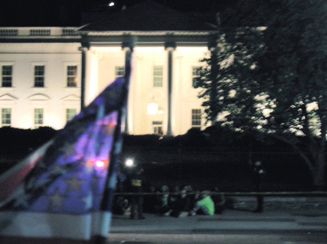 Protestors sit w/their hands cuffed in front of the White House as a plastic flag waves in the foreground.