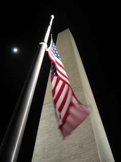 A half moon looks down at  the Washington Monument surrounded by flags at half mast.